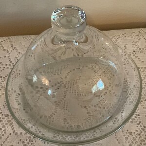 May include: A clear glass cloche with a round base and a domed lid. The cloche has a delicate etched design.