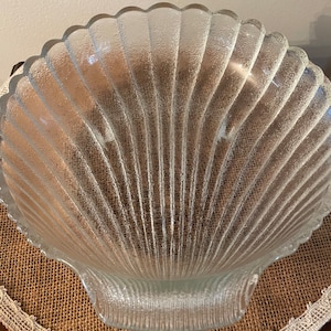 May include: A clear glass serving bowl shaped like a seashell. The bowl has a textured surface and is perfect for serving snacks or fruit.
