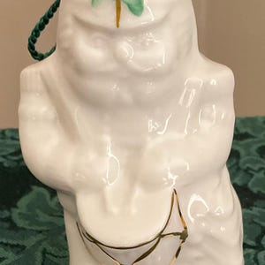 May include: White ceramic figurine of a leprechaun holding a pot of gold. The figurine has a green shamrock painted on the front and a green cord for hanging. The pot of gold has gold accents.