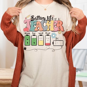 May include: Cream-colored t-shirt with the text "Battery Life of a Teacher" in colorful, retro-style lettering. The design includes battery icons representing the school week, a pencil, an apple, and flowers. A teacher-themed graphic tee.