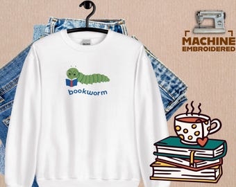 May include: White sweatshirt featuring a green cartoon bookworm reading a book, with the word "bookworm" in blue. The design includes a stack of books, a cup of tea, and a heart. The sweatshirt is hanging on a black hanger.