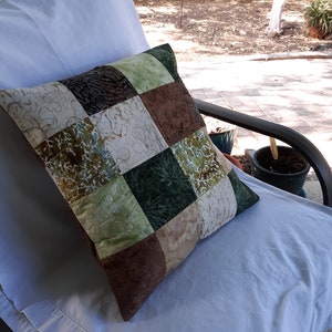 May include: A patchwork pillow with squares of brown, green, and cream fabric. The pillow is sitting on a white chair.