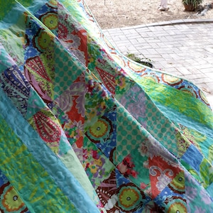 May include: A colorful patchwork quilt with a variety of floral and geometric patterns in shades of blue, green, orange, and pink. The quilt is folded, showcasing the different fabric designs and textures. The quilt appears to be handmade.