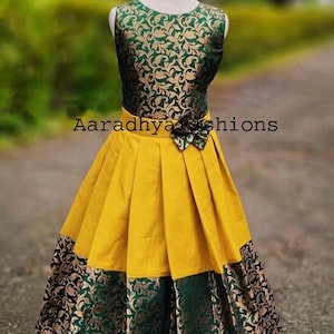 Peut inclure: Robe sans manches avec un corsage &agrave; motifs floraux verts et dor&eacute;s et une jupe pliss&eacute;e jaune. La jupe a une bordure &agrave; motifs verts et dor&eacute;s. Le texte "Aaradhya fashions" est visible sur la robe.