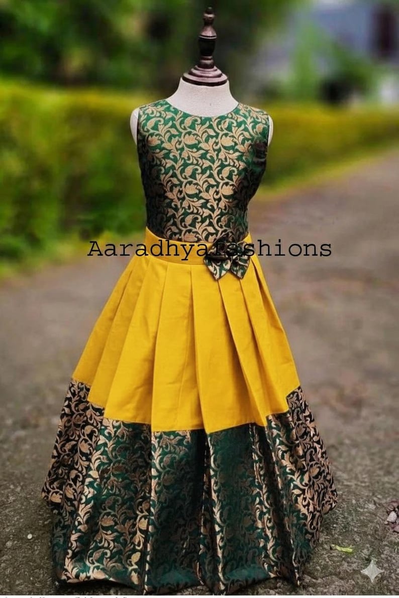 Peut inclure: Robe sans manches avec un corsage &agrave; motifs floraux verts et dor&eacute;s et une jupe pliss&eacute;e jaune. La jupe a une bordure &agrave; motifs verts et dor&eacute;s. Le texte "Aaradhya fashions" est visible sur la robe.