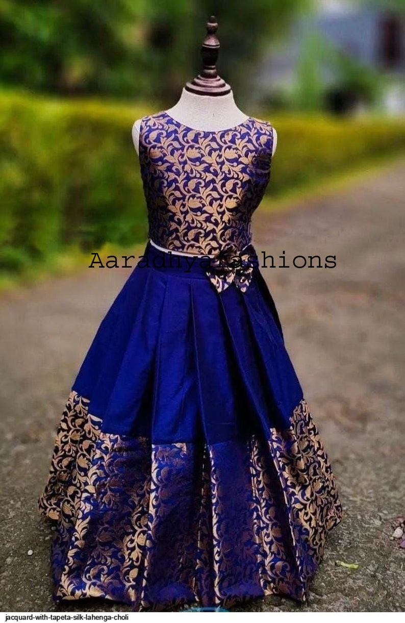 Peut inclure: Une lehenga choli en soie jacquard bleu et or. Le haut sans manches pr&eacute;sente un motif floral dor&eacute; sur fond bleu. La jupe est pliss&eacute;e bleue avec des accents floraux dor&eacute;s. Une ceinture blanche et une fleur ornent l'ensemble.