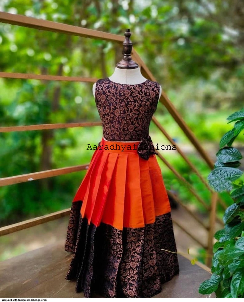 Peut inclure: Robe sans manches sur mannequin. Le corsage est noir avec un motif floral. La jupe est pliss&eacute;e, avec des panneaux orange et noirs. Un n&oelig;ud noir est &agrave; la taille. Le texte "Aaradhyafashions" est visible.