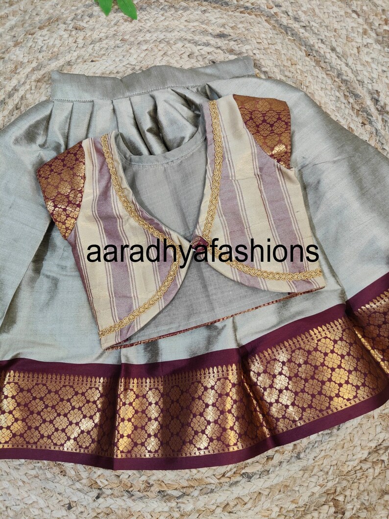 May include: A traditional outfit featuring a silver-gray skirt and a matching top. The vest is striped with gold trim and maroon accents. The skirt has a maroon and gold patterned border. The text "aaradhyafashions" is visible.