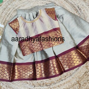 May include: A two-piece outfit featuring a skirt and a blouse. The skirt is a light gray color with a decorative border. The blouse has a striped pattern with gold trim and decorative sleeves. The text "aaradhyafashions" is visible.