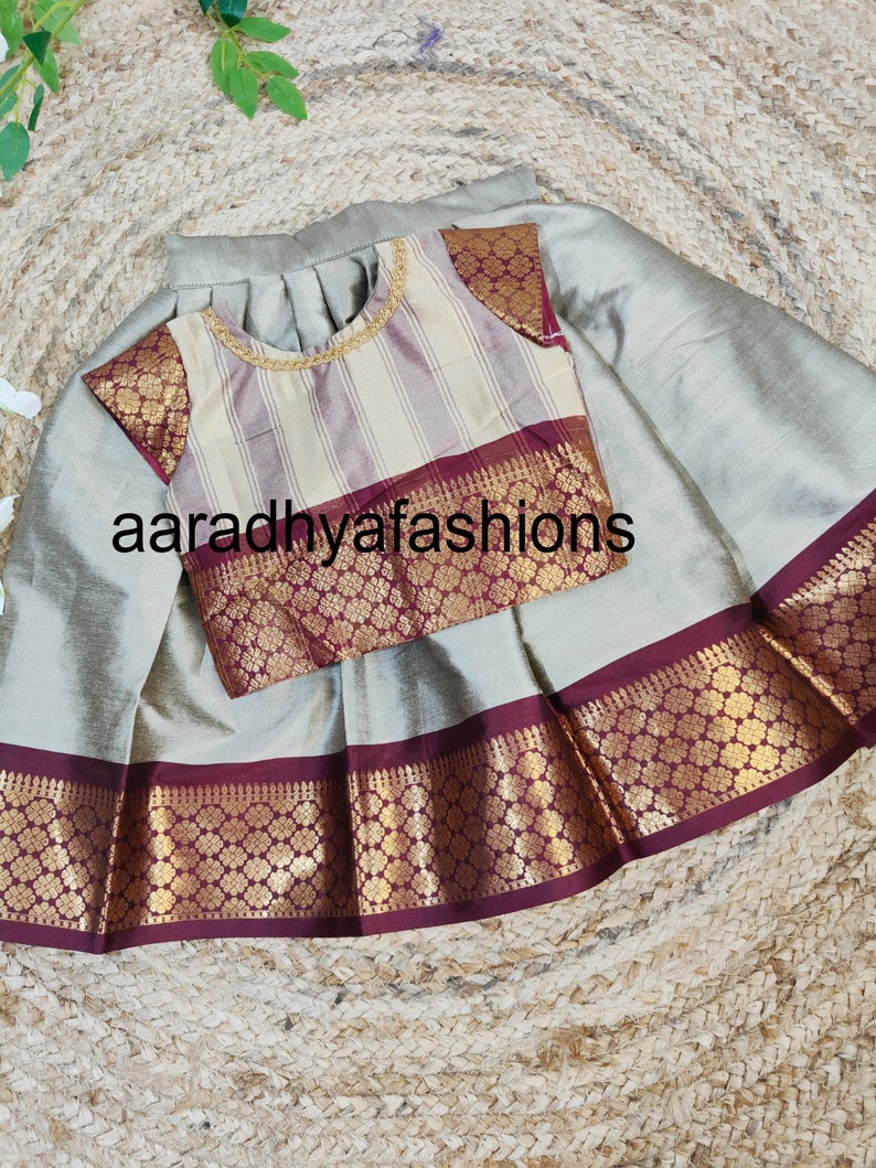 May include: A two-piece outfit featuring a skirt and a blouse. The skirt is a light gray color with a decorative border. The blouse has a striped pattern with gold trim and decorative sleeves. The text "aaradhyafashions" is visible.