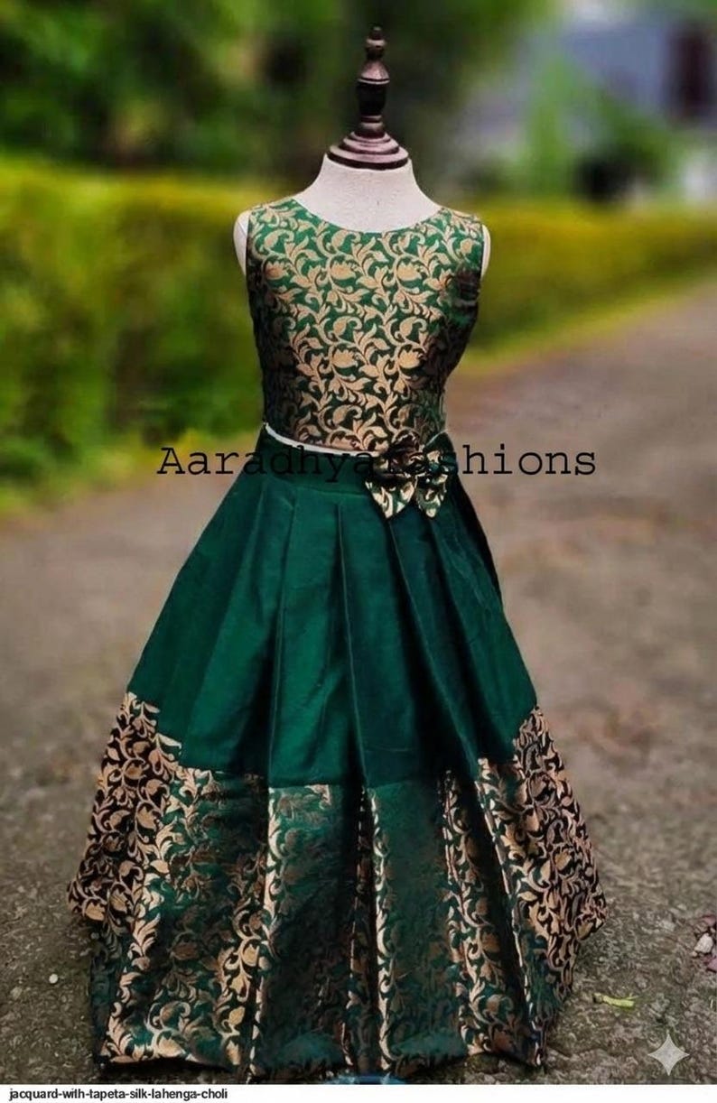 Peut inclure: Une lehenga choli en soie jacquard verte et dor&eacute;e. Le haut sans manches pr&eacute;sente un motif floral dor&eacute; sur fond vert. La jupe est verte avec des panneaux &agrave; motifs floraux dor&eacute;s. Le texte "Aaradhya Fashions" est visible.
