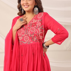 May include: A bright fuchsia dress with detailed embroidery around the V-neck. The dress has three-quarter sleeves and a flowing skirt. Silver jewellery, including earrings and a bracelet, complements the outfit. The dress is a traditional style.