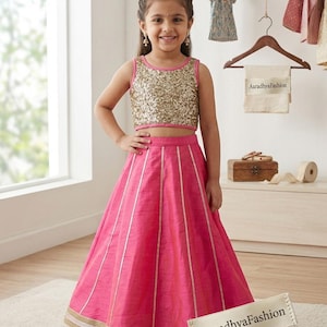 Puede incluir: Una niña con un atuendo tradicional rosa y dorado. La parte superior está cubierta de lentejuelas doradas, y la falda es de un rosa brillante con rayas doradas verticales. El atuendo es de AaradhyaFashion.