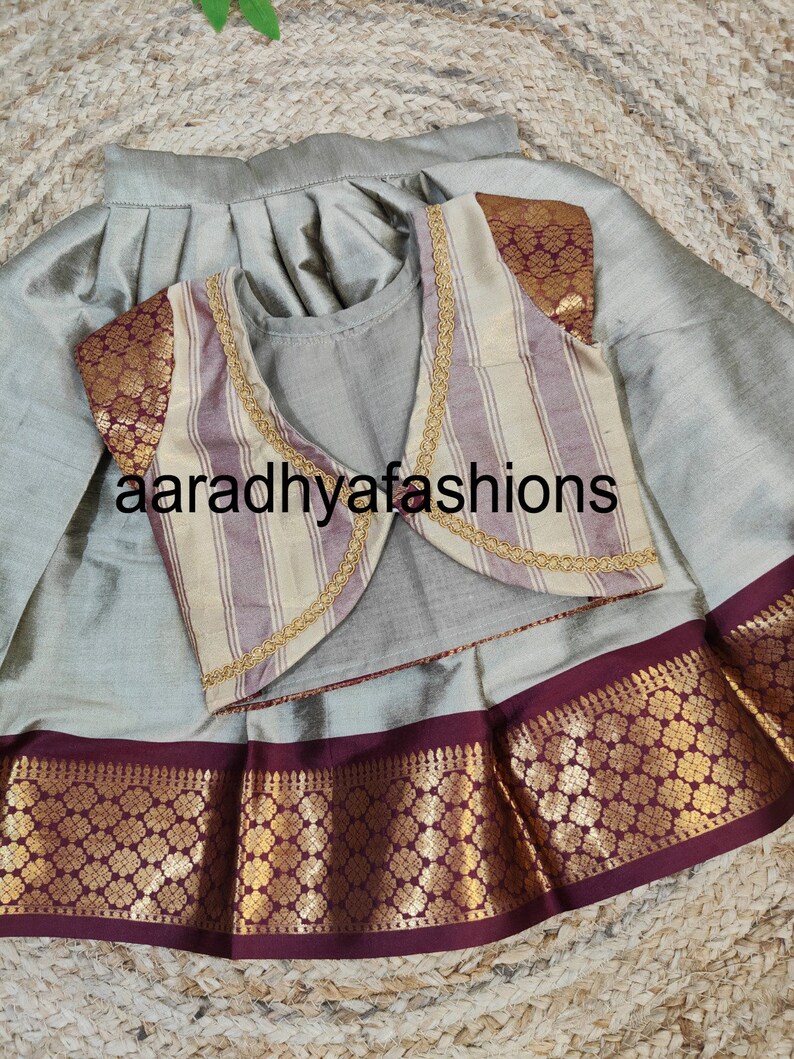 May include: A silver-gray traditional outfit featuring a pleated skirt and a matching top. The outfit includes a striped vest with gold trim and maroon accents. The text "aaradhyafashions" is visible.