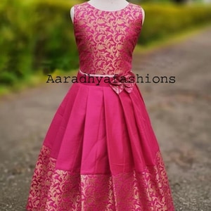 Peut inclure: Robe sans manches fuchsia avec un motif floral dor&eacute; sur le corsage et l'ourlet. La robe pr&eacute;sente une jupe pliss&eacute;e et un n&oelig;ud &agrave; la taille. Le texte "Aaradhyashions" est visible sur la robe.