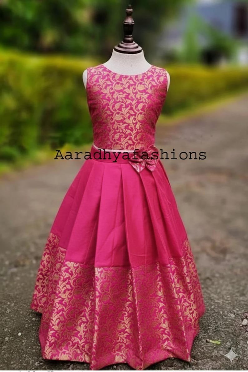 Peut inclure: Robe sans manches fuchsia avec un motif floral dor&eacute; sur le corsage et l'ourlet. La robe pr&eacute;sente une jupe pliss&eacute;e et un n&oelig;ud &agrave; la taille. Le texte "Aaradhyashions" est visible sur la robe.