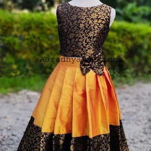 Peut inclure: Robe sans manches avec un corsage &agrave; motifs floraux noirs et dor&eacute;s et une jupe pliss&eacute;e jaune dor&eacute;. La jupe a une bordure &agrave; motifs floraux noirs. La robe est pr&eacute;sent&eacute;e sur un mannequin. Le texte "Aaradhya Fashions" est visible.