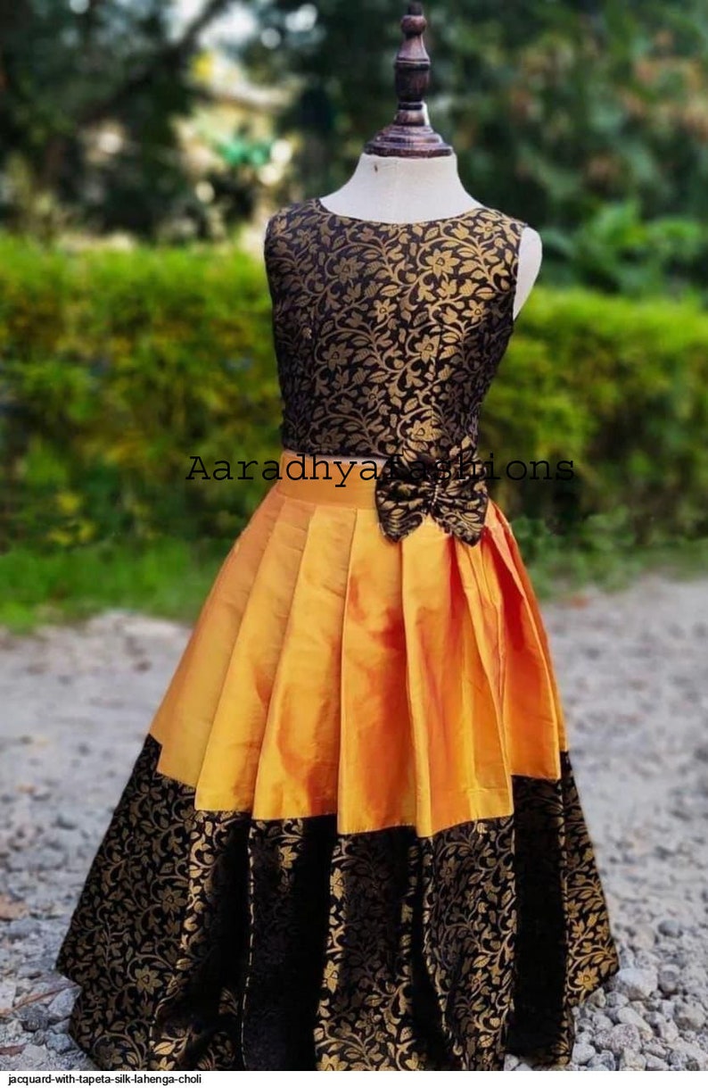 Peut inclure: Robe sans manches avec un corsage &agrave; motifs floraux noirs et dor&eacute;s et une jupe pliss&eacute;e jaune dor&eacute;. La jupe a une bordure &agrave; motifs floraux noirs. La robe est pr&eacute;sent&eacute;e sur un mannequin. Le texte "Aaradhya Fashions" est visible.
