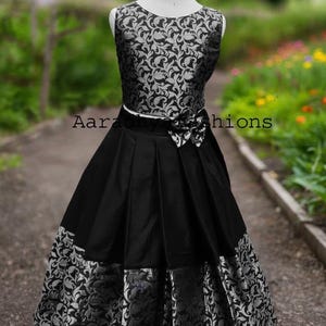 Peut inclure: Robe noire sans manches avec un corsage &agrave; motifs floraux argent&eacute;s et une jupe pliss&eacute;e noire. La jupe a un motif floral argent&eacute; &agrave; l'ourlet. Un n&oelig;ud blanc est &agrave; la taille. Le texte "Aaradhiy hions" est visible.