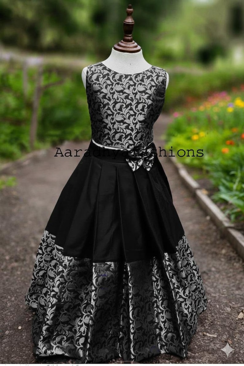 Peut inclure: Robe noire sans manches avec un corsage &agrave; motifs floraux argent&eacute;s et une jupe pliss&eacute;e noire. La jupe a un motif floral argent&eacute; &agrave; l'ourlet. Un n&oelig;ud blanc est &agrave; la taille. Le texte "Aaradhiy hions" est visible.