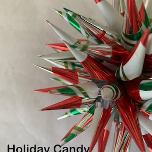 Puede incluir: Un adorno navideño en forma de estrella con detalles de rayas de caramelo rojas, blancas y verdes. El adorno tiene un diseño puntiagudo con conos de papel de aluminio metálico. El texto "Holiday Candy Stripe" se muestra en la parte inferior.
