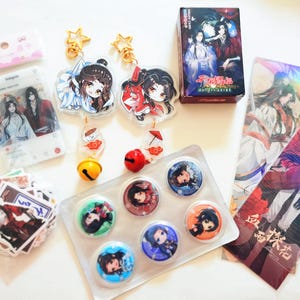 May include: A selection of anime-themed merchandise, including keyrings, stickers, badges, and a small box. The keyrings feature cartoon characters with gold star-shaped clasps. The badges have colourful character designs. The items are arranged on a white surface.