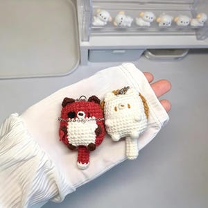 Puede incluir: Dos pequeños llaveros de animales hechos a mano en ganchillo. Uno es una figura de zorro rojo y blanco con un parche negro en el ojo y una cadena plateada. El otro es una figura blanca de gato con orejas pequeñas y una cola.