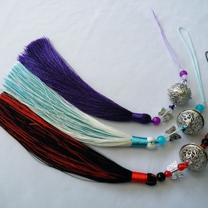 May include: Three decorative tassels in purple, turquoise, and red-black. Each tassel has a beaded accent and a silver bell charm with butterfly details. The tassels are on a white background.