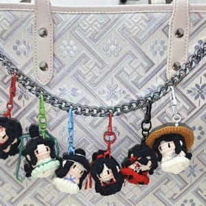 May include: A light-colored tote bag with a silver chain and several crocheted doll keychains. The keychains feature black hair, white skin, and colorful outfits. The bag has a geometric pattern.