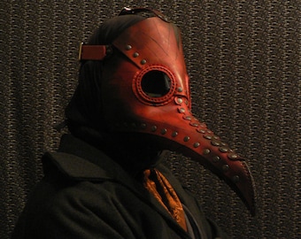 Könnte beinhalten: Eine Person trägt einen schwarzen Mantel und eine rote Ledermaske eines Pestarztes mit einem langen Schnabel. Die Maske hat metallene Akzente und Nieten.