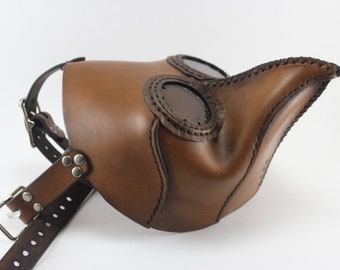 May include: A brown leather plague doctor mask with a long beak and two round eyeholes. The mask has a stitched detail around the edges and is attached to a brown leather strap.