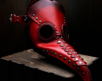 Könnte beinhalten: Eine rote Ledermaske eines Pestarztes mit einem langen Schnabel und Augenlöchern. Die Maske ist mit silbernen Nieten verziert und hat einen Riemen zum Tragen.