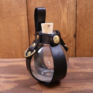 May include: A clear glass bottle with a cork stopper, encased in a black leather holder with brass accents. The holder has a strap for carrying.