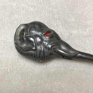 May include: A silver-toned spoon with an elephant head design. The elephant has a detailed trunk, ears, and a red gem for an eye. The spoon is likely made of metal and has a long, slender handle.