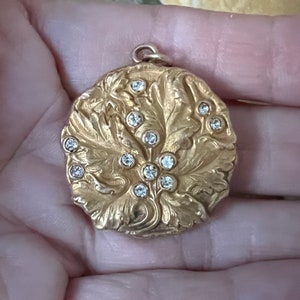 May include: A gold-colored, round pendant with a floral design and embedded clear gemstones. The pendant has a detailed, embossed pattern of leaves and vines, with the gemstones arranged in a cluster. The pendant has a small loop at the top for a chain.