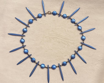 May include: A blue necklace with a circular design. The necklace features a series of blue beads and elongated, pointed blue accents. The necklace is set against a neutral background.