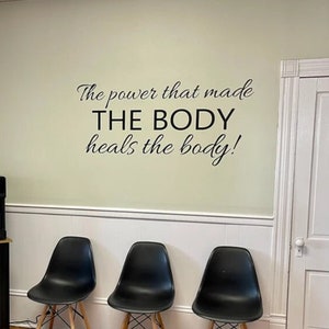 May include: A waiting area with three black chairs with wooden legs, a black side table with a water cooler, and a quote on the wall that reads 'The power that made THE BODY heals the body!'