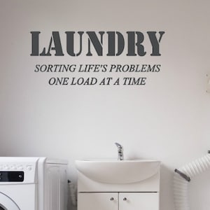 Op de afbeelding: Een witte wasruimte met twee wasmachines, een witte gootsteen en een witte kast. De muur heeft een bord met de tekst "Laundry, Sorting Life's Problems One Load At A Time."