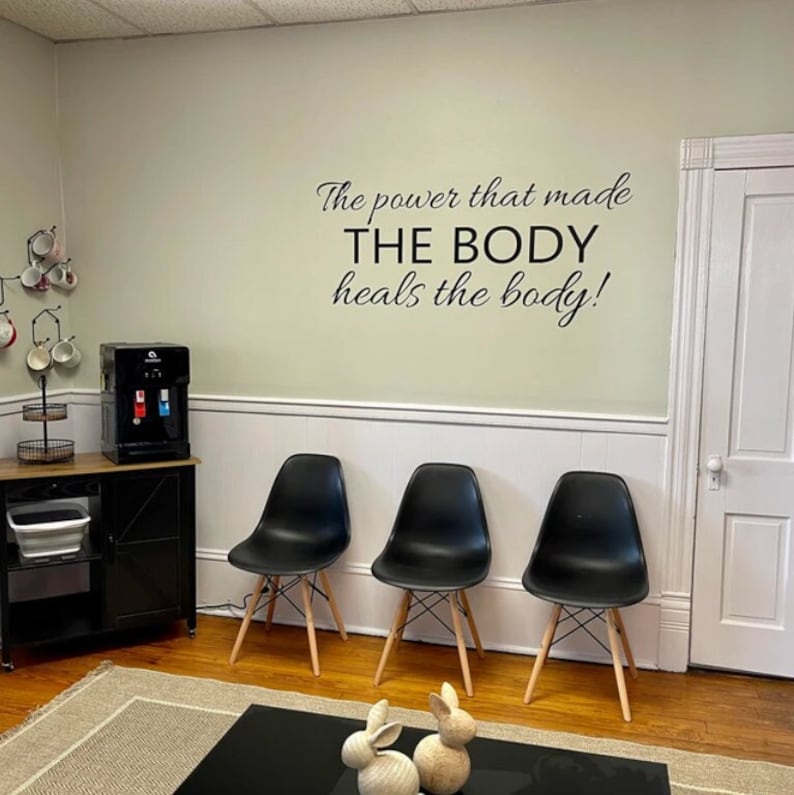 May include: A waiting area with three black chairs with wooden legs, a black side table with a water cooler, and a quote on the wall that reads 'The power that made THE BODY heals the body!'