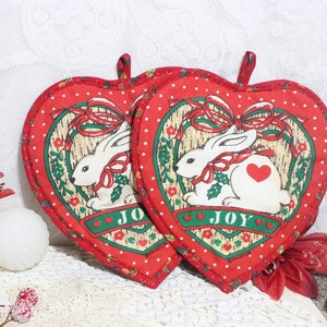 May include: Two heart-shaped red pot holders with white polka dots and a white rabbit design. The design includes a red bow, green holly, and the word "JOY". A small loop is at the top for hanging.