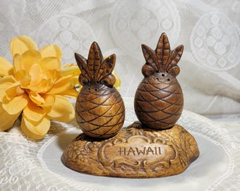 May include: A wooden salt and pepper shaker set shaped like pineapples with a carved wooden base that says "HAWAII".