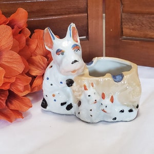 May include: A ceramic planter in the shape of a dog with puppies. The white planter has black spots and orange and blue accents. The planter is on a white surface with orange flowers in the background.