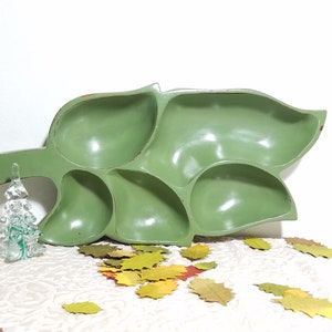 May include: A green leaf-shaped serving tray with multiple compartments. A small glass Christmas tree ornament is to the left of the tray. Scattered around the tray are decorative leaf cutouts in various fall colors.