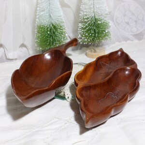 May include: Two wooden leaf-shaped serving dishes. One has a handle, the other has two compartments with carved floral designs. The dishes are a rich brown color and sit on a white surface with small Christmas trees in the background.