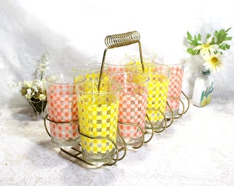 May include: A set of six vintage drinking glasses in a metal carrier. The glasses feature a checkered pattern in pink and yellow. The metal carrier has a gold finish and a handle.