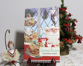 May include: A vintage Christmas Recipes booklet on a wooden stand. The cover features a festive illustration of a holiday table with food and a snowy window scene. A small Christmas tree and a doll ornament are also in the image.