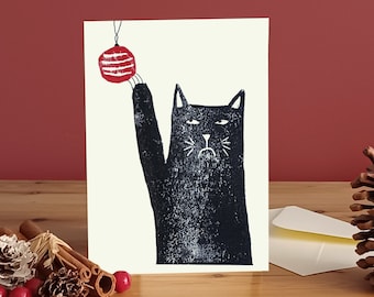 May include: A black cat with a grumpy expression reaches up to a red and white Christmas ornament hanging from a string. The cat is printed on a white card.