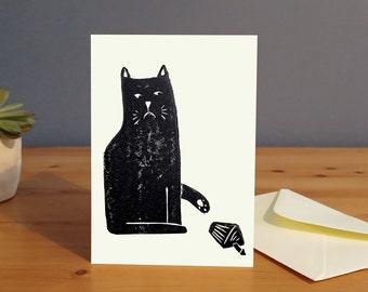 May include: A black and white greeting card featuring a black cat with a grumpy expression. The cat is sitting and looking at a small, white cupcake on the ground.
