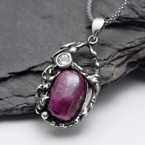 May include: A silver pendant necklace with a large, oval-shaped, pink gemstone set in a silver frame. A smaller, clear gemstone is set above the larger gemstone.