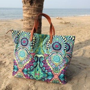 May include: A large, colorful tote bag with a paisley pattern in shades of blue, green, pink, and yellow. The bag has brown leather handles and is sitting on a sandy beach.